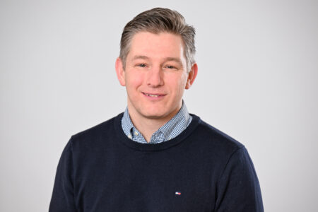 Porträt einer Person mit kurz gescheitelten, dunkelgrauen Haaren, die ein dunkelblaues Oberteil über einem blau-weiß karierten Hemd trägt und vor hellem, neutralem Hintergrund in die Kamera blickt.