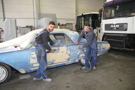 Zwei Personen in Arbeitskleidung schleifen mit Elektroschleifern die teils abgebeizte Karosserie eines hellblauen Oldtimers in einer Werkstatt; im Hintergrund stehen Lkw.