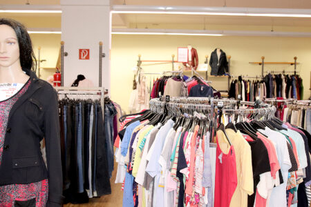 Blick in ein Bekleidungsgeschäft mit einer Schaufensterpuppe links und einem runden Kleiderständer voller bunter T‑Shirts im Vordergrund, dahinter weitere Ständer mit Hosen und Jacken.