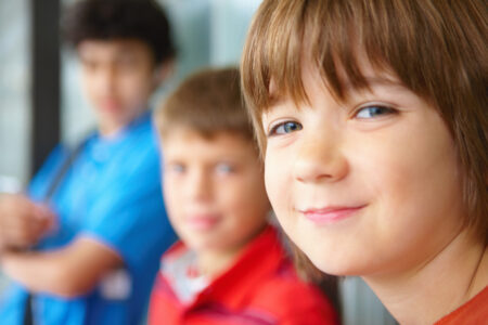 Nahaufnahme eines Kindes mit braunen Haaren, das in die Kamera blickt; im Hintergrund sind zwei weitere Kinder unscharf zu sehen, eines in rotem und eines in blauem Oberteil.