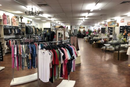 Großer, hell beleuchteter Ladeninnenraum mit Kleiderständern voller bunter Oberteile im Vordergrund, Regalen mit Kleidung an der linken Wand und Tischen mit Geschirr, Vasen und künstlichen Blumen rechts, auf Holzoptikboden unter Deckenleuchten und Kronleuchtern.