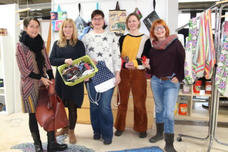 Fünf Personen stehen in einem Raum vor einer Wand mit aufgehängten Taschen und halten u. a. eine Ledertasche, eine grüne Kiste mit Reißverschlüssen, einen gepunkteten Turnbeutel und Scheren; rechts steht ein Kleiderständer mit bunten Stofftaschen.