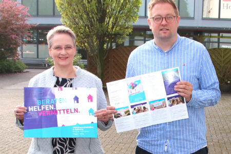 Zwei Personen stehen im Freien vor einem Gebäude; die linke Person hält ein Plakat mit der Aufschrift „BERATEN. HELFEN. VERMITTELN.“, die rechte Person hält eine aufgeklappte Broschüre.