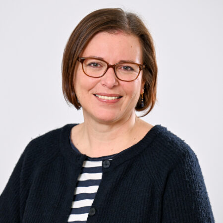 Porträt einer lächelnden Person mit kurzen braunen Haaren und Brille, die ein dunkelblaues Strickcardigan mit Knöpfen über einem schwarz-weiß gestreiften Shirt trägt, vor neutralem Hintergrund.