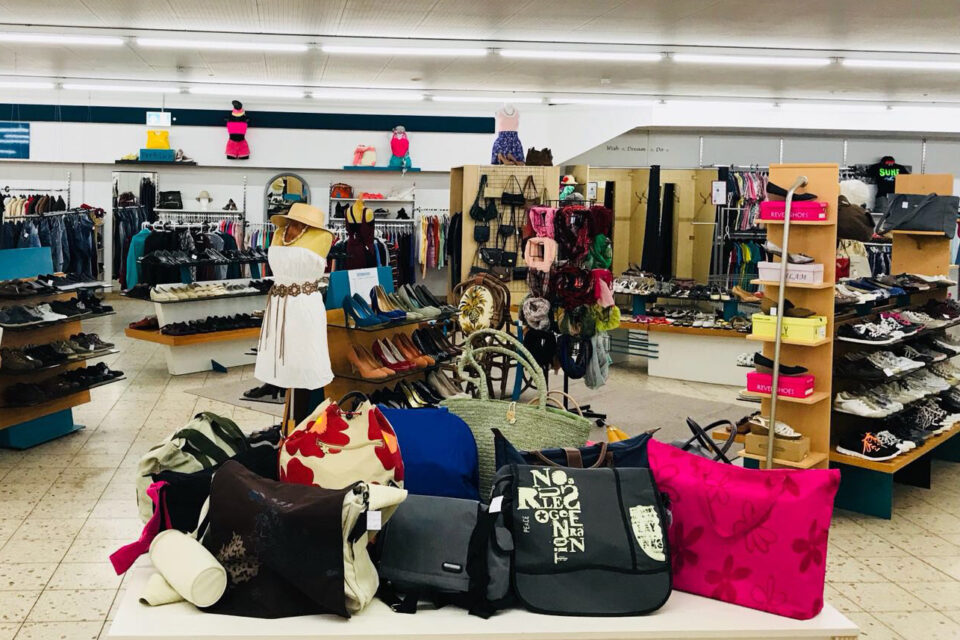 Innenansicht eines Bekleidungsgeschäfts mit vielen Regalen voll Schuhen und Accessoires; vorn ein Tisch mit bunten Taschen, daneben eine Schaufensterpuppe im weißen Kleid, im Hintergrund Kleiderständer mit Kleidung.