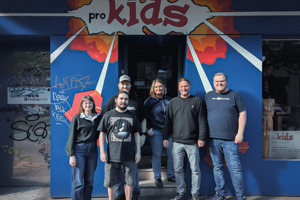 Sechs Personen stehen vor einem blauen Eingangsbereich mit Wandbild, auf dem in einer roten Wolke mit weißen Strahlen „pro kids“ steht; links ist eine verglaste Tür mit Graffiti und dem Schild „diakoniewerk Duisburg GmbH“ zu sehen.