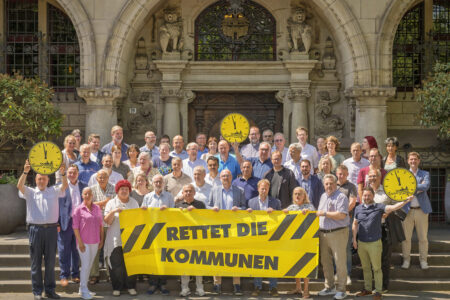Eine große Gruppe Menschen steht auf den Stufen vor einem steinernen Gebäude mit Bögen und Löwenfiguren und hält ein gelbes Banner mit der Aufschrift „RETTET DIE KOMMUNEN“ sowie mehrere gelbe Uhren-Schilder.