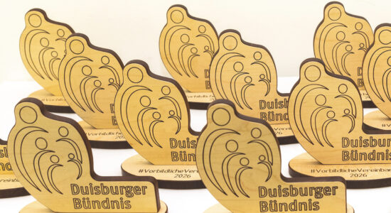 Mehrere gleichförmige Holz-Trophäen mit eingravierten Linien und Kreisen stehen auf einem weißen Tisch und tragen die Aufschrift „Duisburger Bündnis“ sowie „#VorbildlicheVereinbarkeit 2026“.