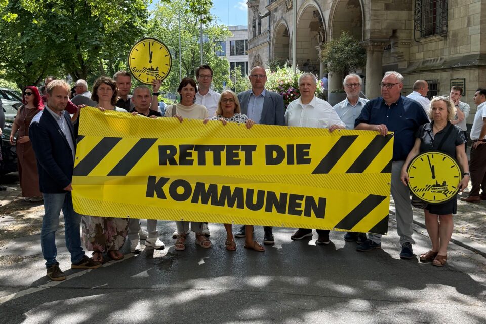 Eine Gruppe von rund 15 Menschen steht auf einer Straße vor einem alten steinernen Gebäude und hält ein großes gelbes Banner mit der Aufschrift „RETTET DIE KOMMUNEN“, rechts und im Hintergrund sind gelbe Uhr-Schilder zu sehen.