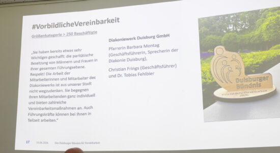 Eine projizierte Präsentationsfolie zeigt die Überschrift #VorbildlicheVereinbarkeit, links einen längeren Textblock und rechts Diakoniewerk Duisburg GmbH mit mehreren Namen sowie ein Foto eines hölzernen Aufstellers mit der Aufschrift Duisburger Bündnis #VorbildlicheVereinbarkeit 2026. Am unteren Bildrand sind zwei angeschnittene Personen zu sehen, eine davon mit Brille.