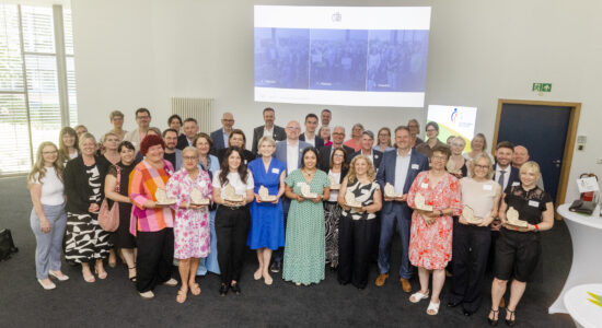 Eine große Gruppe Erwachsener steht in einem hellen Konferenzraum für ein Gruppenfoto; mehrere Personen in der ersten Reihe halten helle Holzplaketten, im Hintergrund sind eine projizierte Folie und ein Aufsteller zu sehen.
