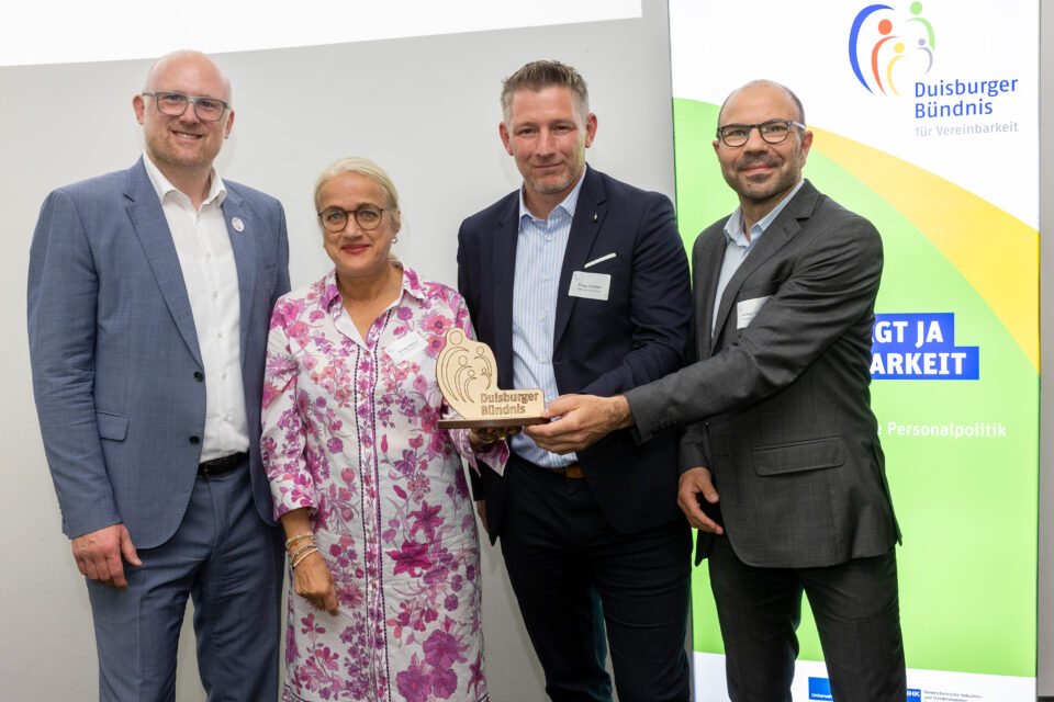 Vier Personen stehen nebeneinander vor einem Roll-up mit der Aufschrift „Duisburger Bündnis für Vereinbarkeit“, wobei die beiden Männer rechts eine kleine Holzplakette mit der Gravur „Duisburger Bündnis“ in die Kamera halten.