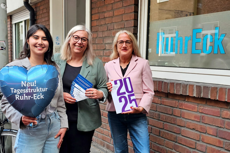 Drei Frauen stehen vor einer Backsteinwand mit dem Fensteraufdruck „RuhrEck“; links hält eine einen blauen herzförmigen Ballon mit der Aufschrift „Neu! Tagesstruktur Ruhr-Eck“, die mittlere mehrere Flyer und die rechte ein Heft mit der Zahl „2025“.