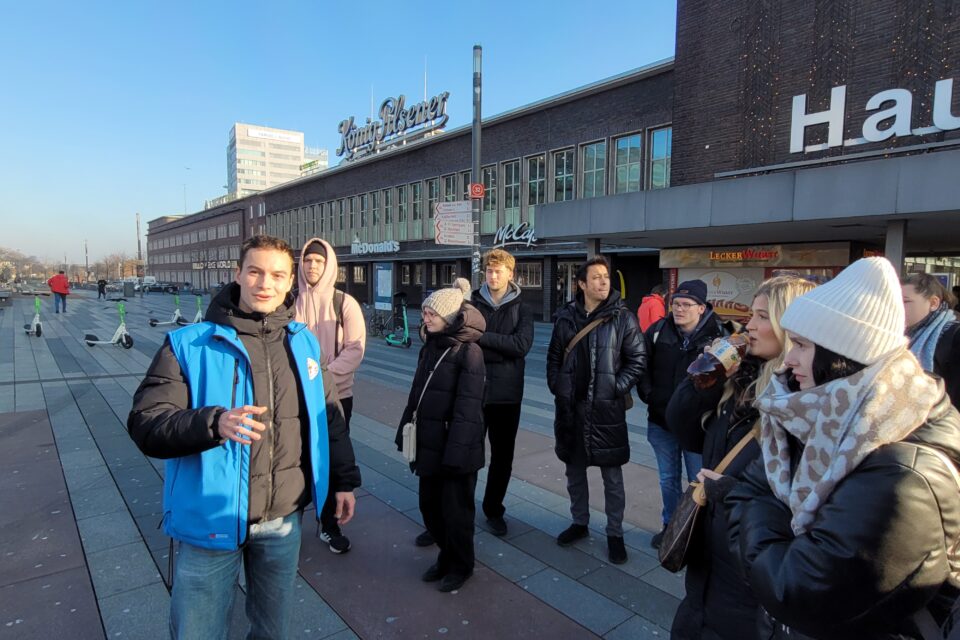 Eine Gruppe von Menschen in Winterkleidung steht auf einem Platz vor einem langen Backsteingebäude mit den Schildern „König Pilsener“, „McDonald’s“ und „Lecker Wurst“, während vorn links ein Mann in blauer Weste in die Kamera blickt; im Hintergrund stehen mehrere E‑Scooter.