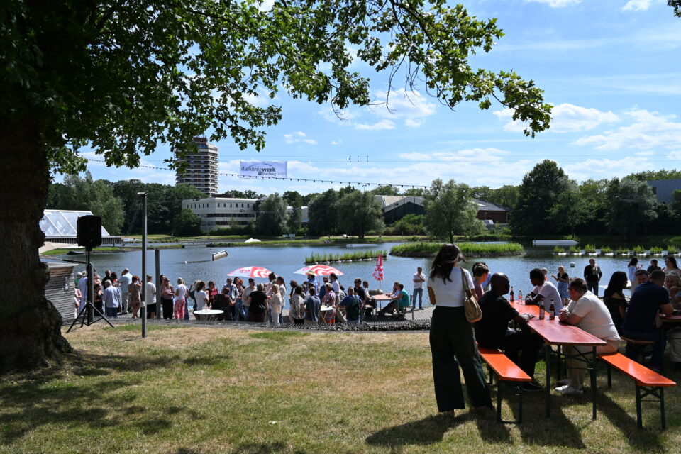Viele Menschen versammeln sich am Ufer eines Sees auf Wiese und Pflaster, sitzen an orangefarbenen Bänken und unter rot-weißen Sonnenschirmen, im Hintergrund Bäume, mehrere Gebäude und ein hoher Turm, darüber eine Lichterkette mit Banner unter blauem Himmel.