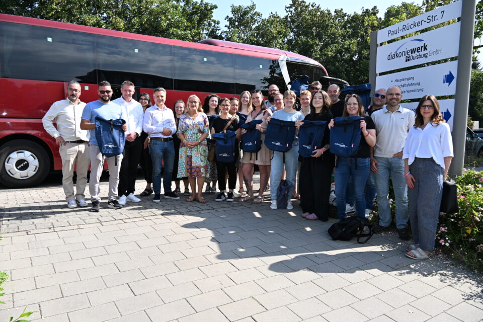 Eine größere Gruppe Menschen steht auf einem Gehweg vor einem roten Reisebus neben einem Schild mit der Aufschrift „diakoniewerk Duisburg GmbH, Paul-Rücker-Str. 7“, mehrere halten dunkelblaue Rucksäcke hoch.