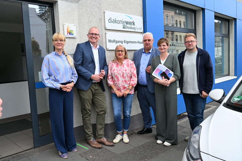 Sechs Erwachsene stehen dicht nebeneinander vor einem Gebäude mit blau abgesetzter Fassade neben Schildern mit der Aufschrift „diakoniewerk Duisburg GmbH“ und „Wolfgang-Eigemann-Haus“, zwei halten Broschüren in der Hand.
