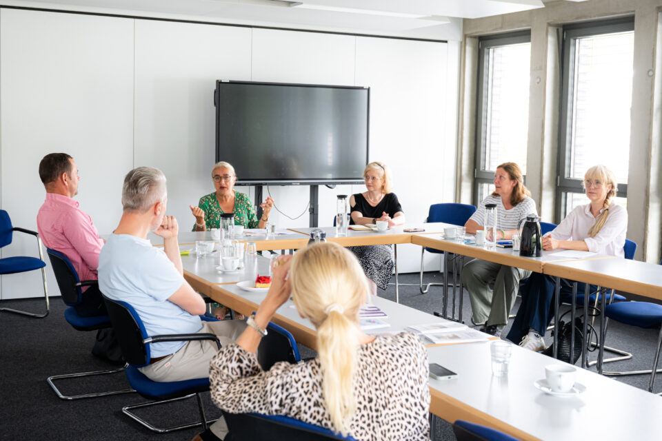 Sieben Personen sitzen in einem hellen Konferenzraum an u-förmig zusammengestellten Tischen; vor einem großen Bildschirm spricht eine Person in grüner Kleidung und gestikuliert, während auf den Tischen Tassen, Gläser, Karaffen, Papiere und eine Thermoskanne stehen.