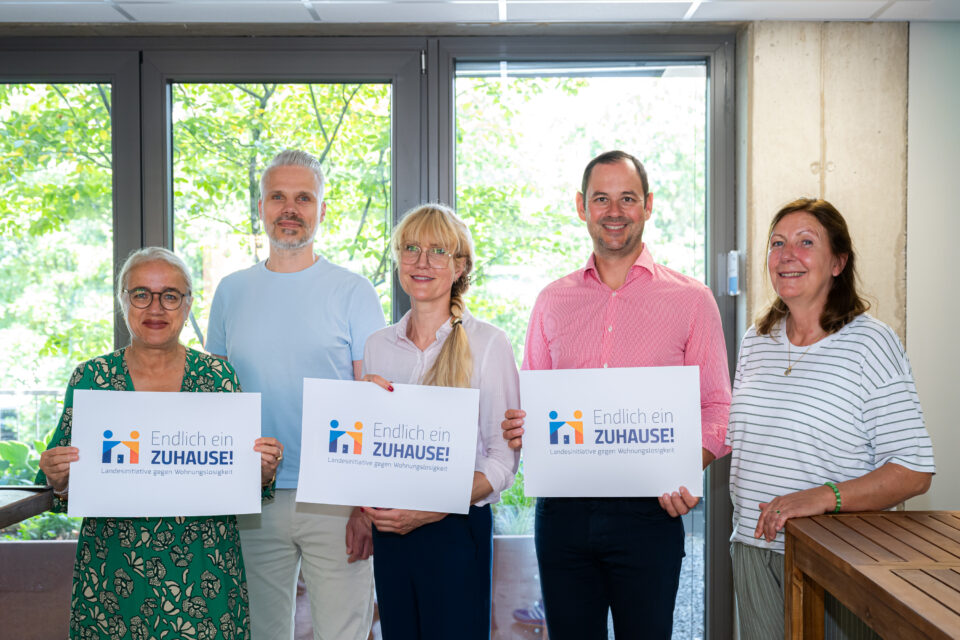 Fünf Erwachsene stehen in einem Raum vor großen Fenstern mit grünem Laub draußen; drei von ihnen halten Plakate mit der Aufschrift „Endlich ein ZUHAUSE! Landesinitiative gegen Wohnungslosigkeit“.