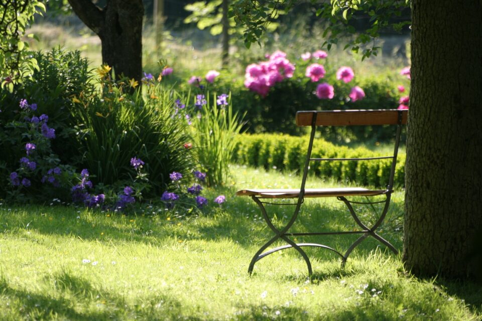 Ein Gartenstuhl aus Holz und Metall steht auf einer Rasenfläche im Schatten eines Baums. Dahinter liegen Beete mit rosa und lila Blumen sowie einer niedrigen Hecke.