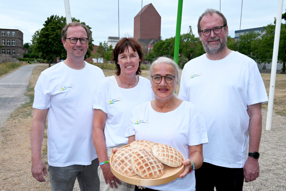 Vier Erwachsene in weißen T‑Shirts stehen im Freien; vorne hält eine Person mit Brille ein rundes Holztablett mit mehreren hellen Fladenbroten. Im Hintergrund sind Bäume, ein Kiesweg und städtische Gebäude zu sehen.