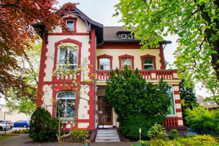 Mehrstöckiges cremefarbenes Haus mit roten Zierrahmen, Balkon mit Balustrade und Eingangstreppe (Hausnummer 155), umgeben von dichtem Grün und Bäumen; links am Bildrand stehen geparkte Autos.