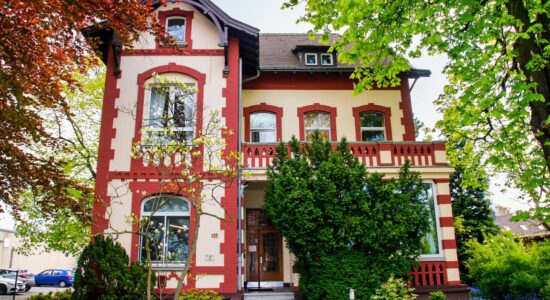 Mehrstöckiges cremefarbenes Haus mit roten Zierrahmen, Balkon mit Balustrade und Eingangstreppe (Hausnummer 155), umgeben von dichtem Grün und Bäumen; links am Bildrand stehen geparkte Autos.