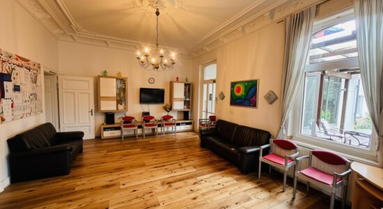 Heller Raum mit Stuckdecke und Kronleuchter, Holzdielenboden, zwei schwarzen Ledersofas, mehreren rot-grauen Stühlen, Wandregalen mit Fernseher und Uhr, großen Fenstern mit Vorhängen und Blick auf eine überdachte Terrasse mit Tischen und Stühlen; an den Wänden hängen bunte Bilder.