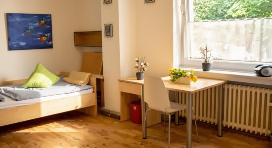 Helles Zimmer mit Holzfußboden: links ein Einzelbett mit grauer Bettwäsche und grünem Kissen, rechts ein Schreibtisch mit weißem Stuhl, Topfpflanzen und gelben Blumen am Fensterbrett über einem Heizkörper; an der Wand hängen Bilder, und auf dem Fensterbrett liegt ein Kopfhörer.