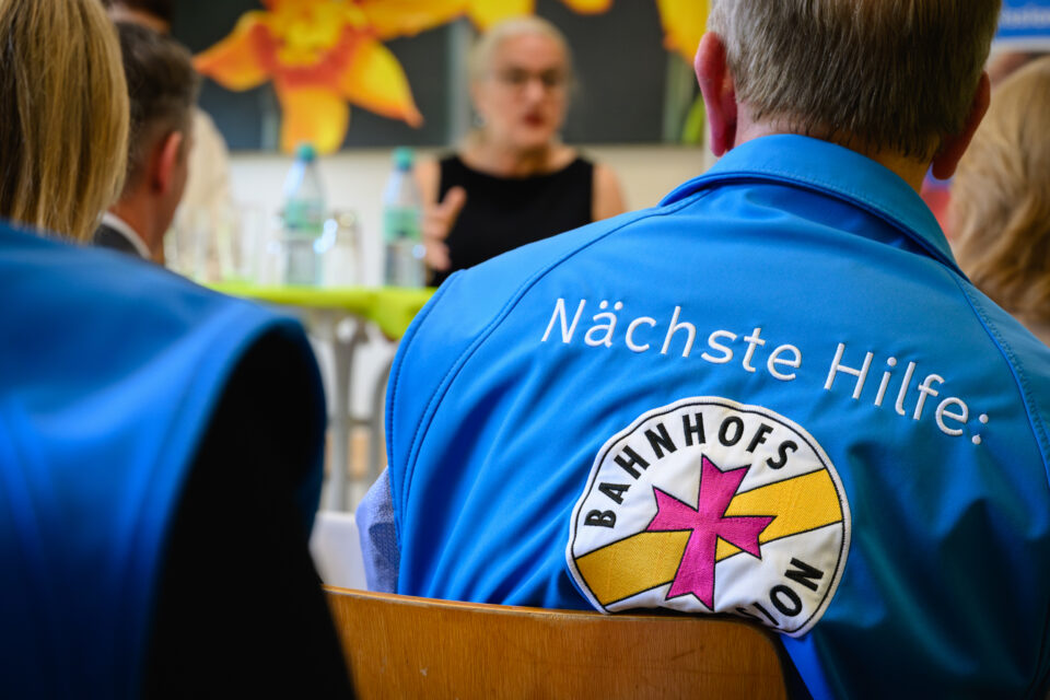 Rückenansicht einer Person in blauer Weste mit der Aufschrift „Nächste Hilfe:“ und dem Logo „Bahnhofsmission“; im Hintergrund unscharf eine sprechende Person, Wasserflaschen auf einem Tisch und ein großes Blumenbild an der Wand.