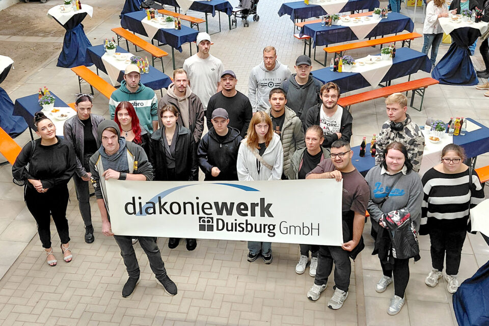 Etwa 20 junge Menschen stehen dicht beisammen und halten ein großes Banner mit der Aufschrift „diakoniewerk Duisburg GmbH“, umgeben von Reihen gedeckter Tische und Bänke mit blauen Tischdecken, Getränkeflaschen und kleinen Pflanzendekorationen.