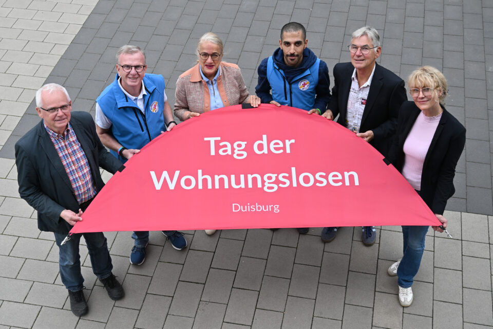 Sechs Erwachsene stehen auf grauem Pflaster und halten gemeinsam ein großes, halbkreisförmiges rotes Banner mit der Aufschrift „Tag der Wohnungslosen“ und darunter „Duisburg“.