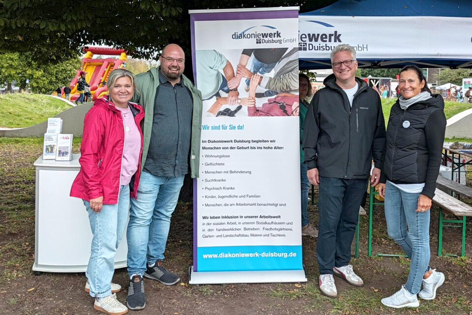 Vier Erwachsene stehen im Freien neben einem Roll-up-Banner „diakoniewerk Duisburg GmbH“ und einem kleinen Infostand unter einem blauen Pavillon; im Hintergrund ist eine rote aufblasbare Rutsche mit spielenden Kindern zu sehen.
