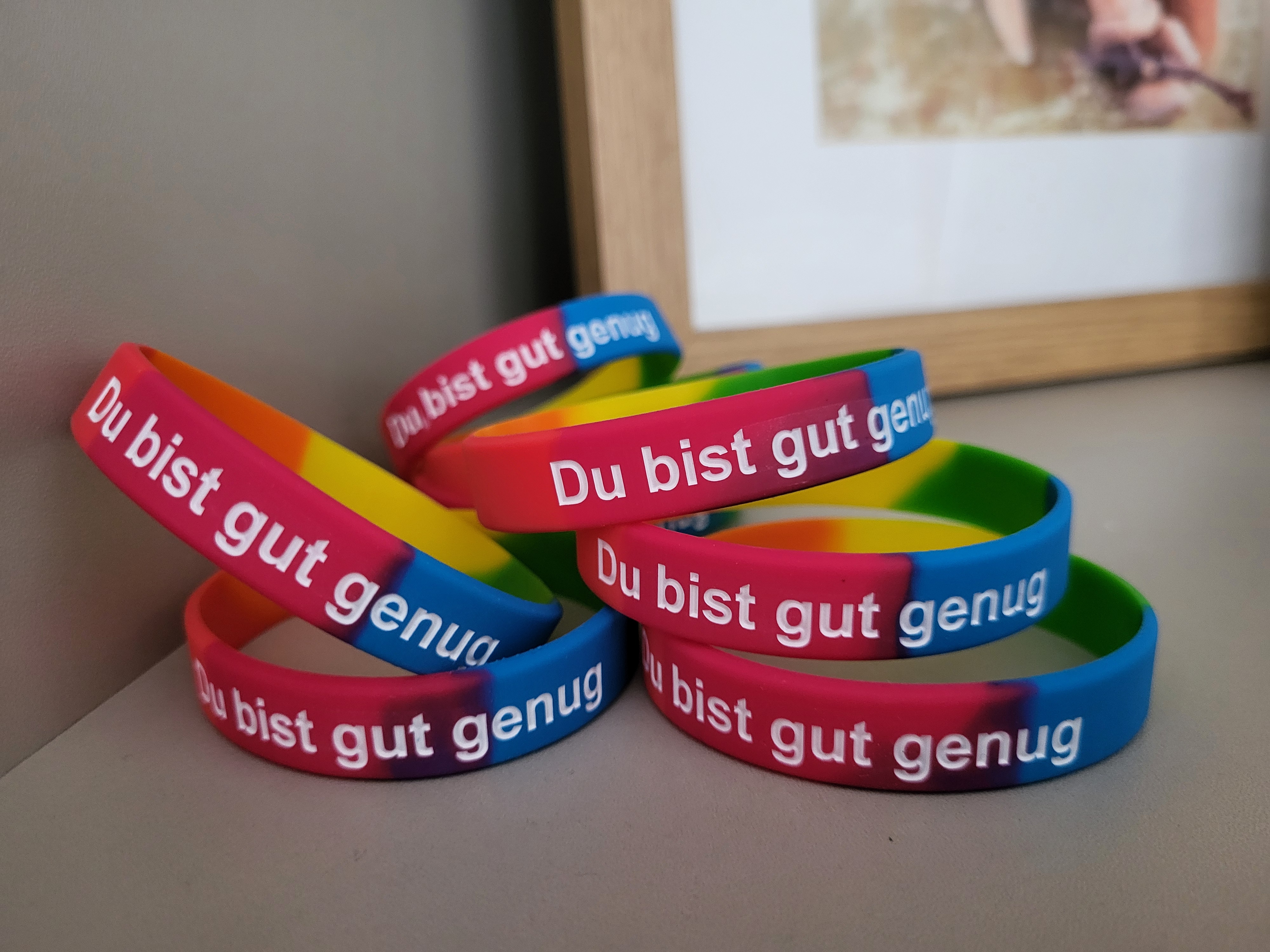 Mehrere bunte Silikonarmbänder mit der weißen Aufschrift „Du bist gut genug“ liegen gestapelt auf einer Oberfläche neben einem Holz-Bilderrahmen.